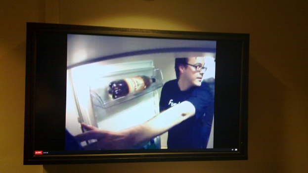 Ein Fernseher, der an einer Wand montiert ist und eine Person, einen Kühlschrank und eine Flasche auf einem Kühlschrankregal zeigt.