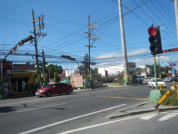 Roter Wagen fährt auf einer Straße mit einer Ampel, Strommasten und Drähten an Gebäuden, Bäumen und einem klaren blauen Himmel im Hintergrund.